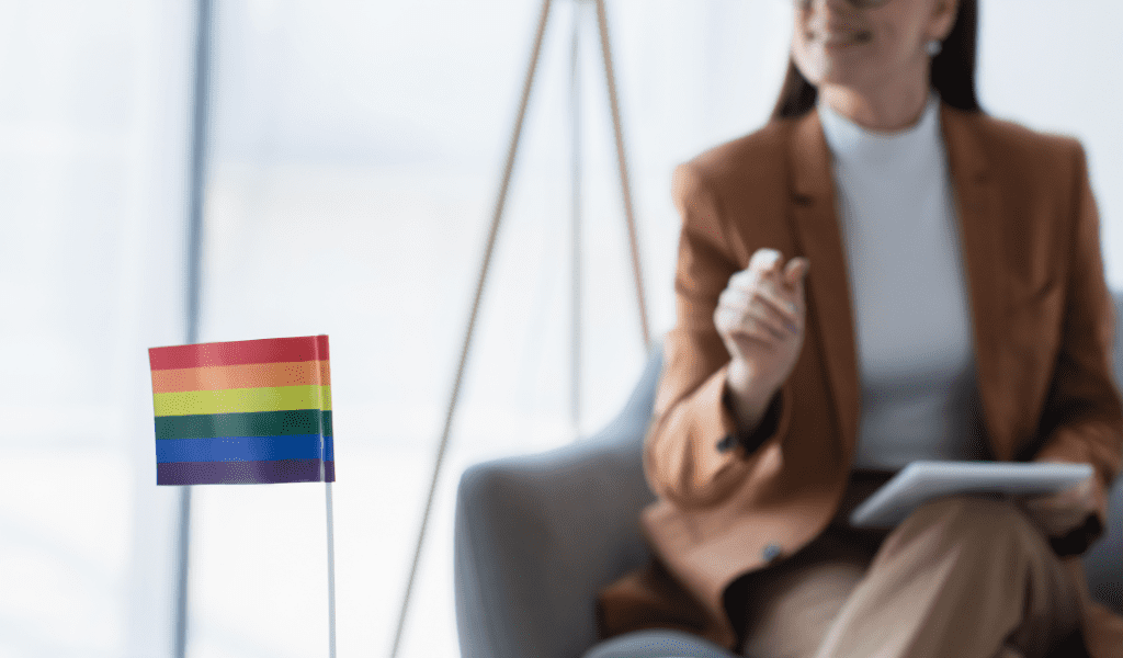 Woman sitting in chair at work with LGBTQ Pride flag on table in front of her.